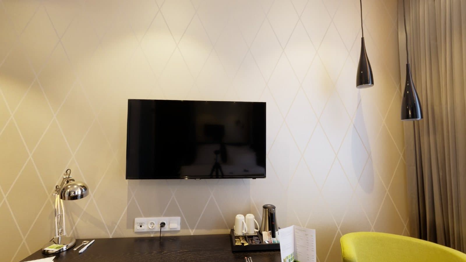 A hotel room wall with a television, a desk and a lamp